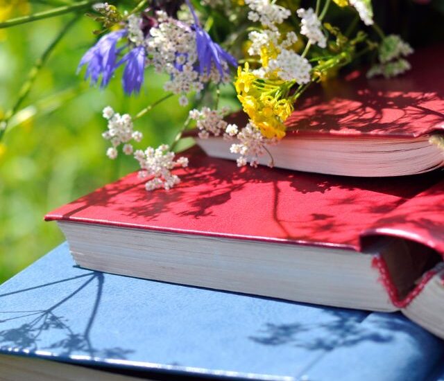 Pile of books with bright covers in red, blue, and teal laying on grass and small spring flowers in white, yellow, and purple on top